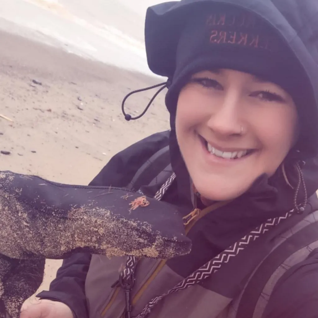 Elizabeth Forbes, wearing a coat with her hood up and a hat underneath, smiling at the camera and holding a prosthetic leg upside down. She is stood on the beach. 