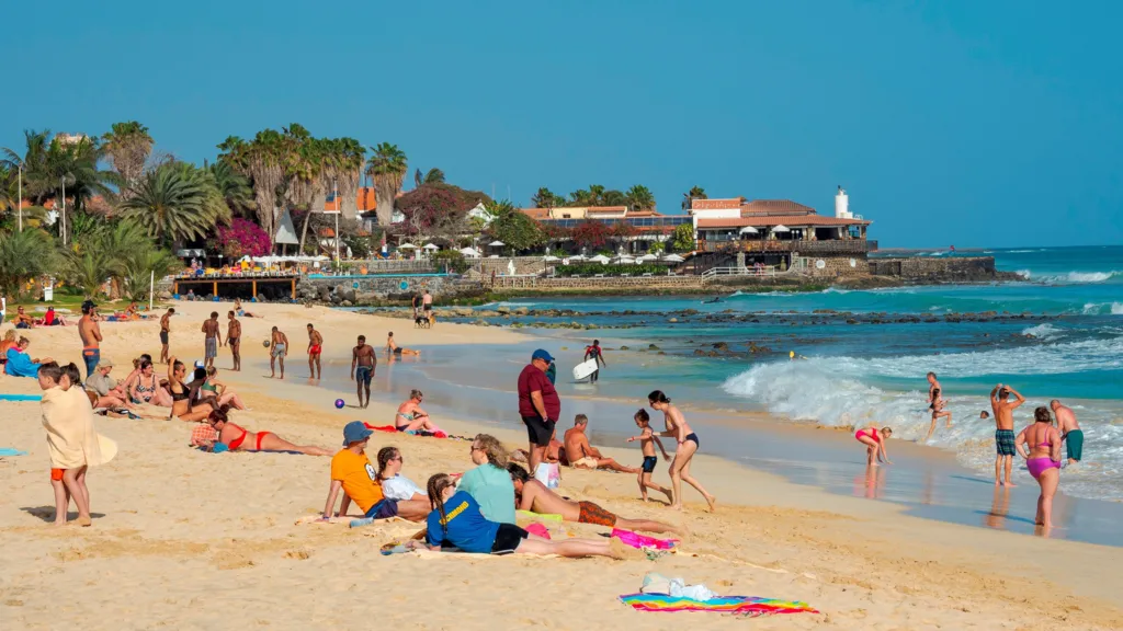 Holidaymakers relax on a beach on the island of Sal in Cape Verde. Many are lying or sitting on towels on the sand, while others are standing in or near the sea.
