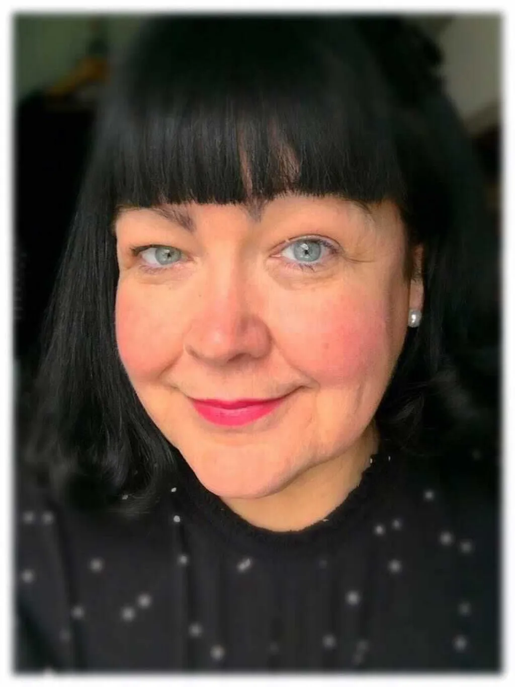 A woman with black hair and pearl earrings is looking into the camera. She is wearing a black blouse with white dots. 
