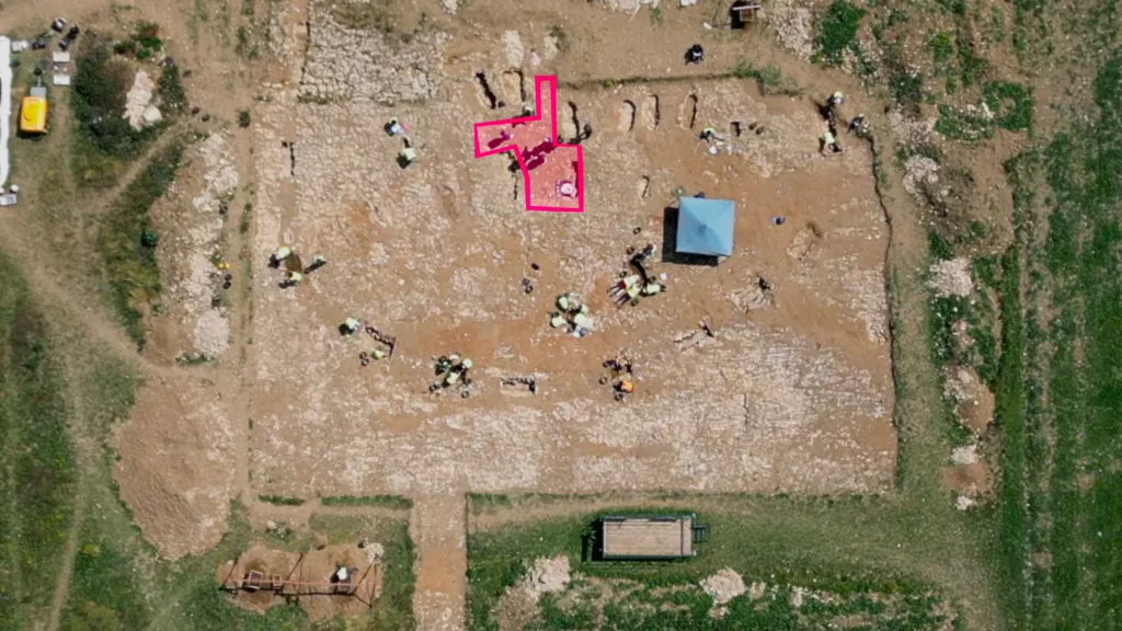 Aerial view of the archaeological dig site. The location of a possible building has been graphically highlighted in pink. Six or seven graves are clustered around it.  There are another six or seven graves visible that are located further from the highlighted feature.  Archaeologists are working at different parts of the site wearing yellow high visibility jackets. The wider area of the cemetery that is being excavated has exposed brown soil. It is surrounded by a green field. 