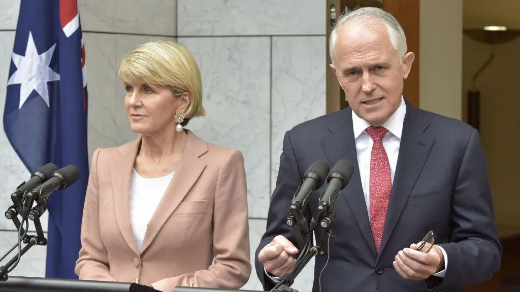 A woman, with short blond hair and in a light suit, and a man in a dark suit, both standing at a podium with a partial Australian flag in background.