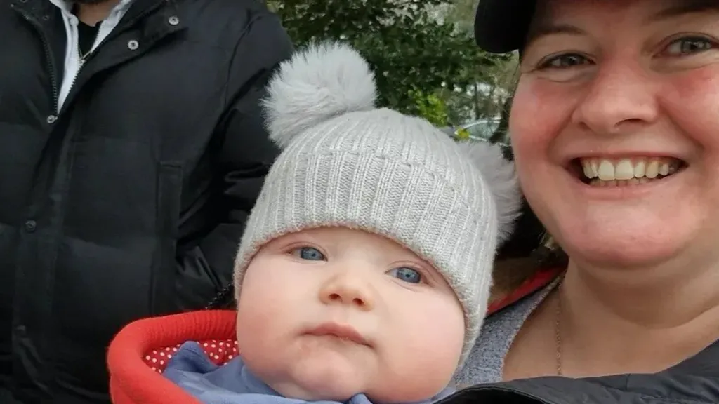 Emma is smiling and holding Theo in front of her - he's wearing a grey hat and red jumper