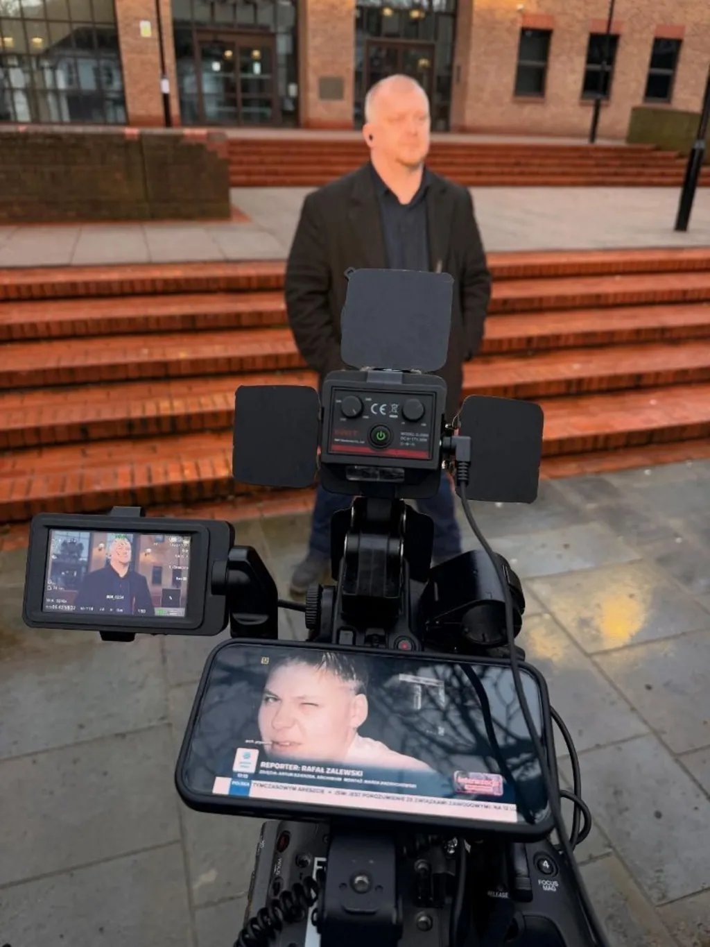 A man in front of a camera on the steps of a crown court. A woman's face can be seen on a phone screen in the foreground.