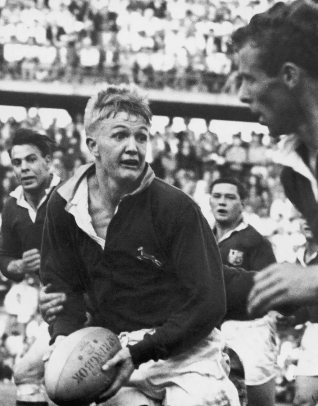 A black and white photograph of a rugby match. One man is running with the ball, being pursued by three others. 