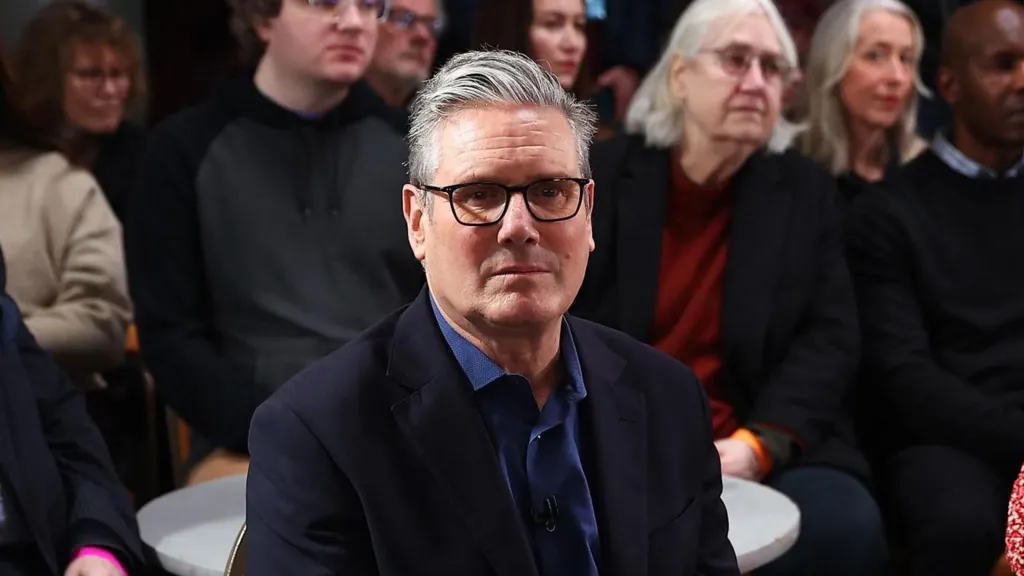 Keir Starmer, wearing black rimmed glasses and an open necked blue shirt, with suit jacket, stares plaintively at the camera. He is sitting in the audience of a press event