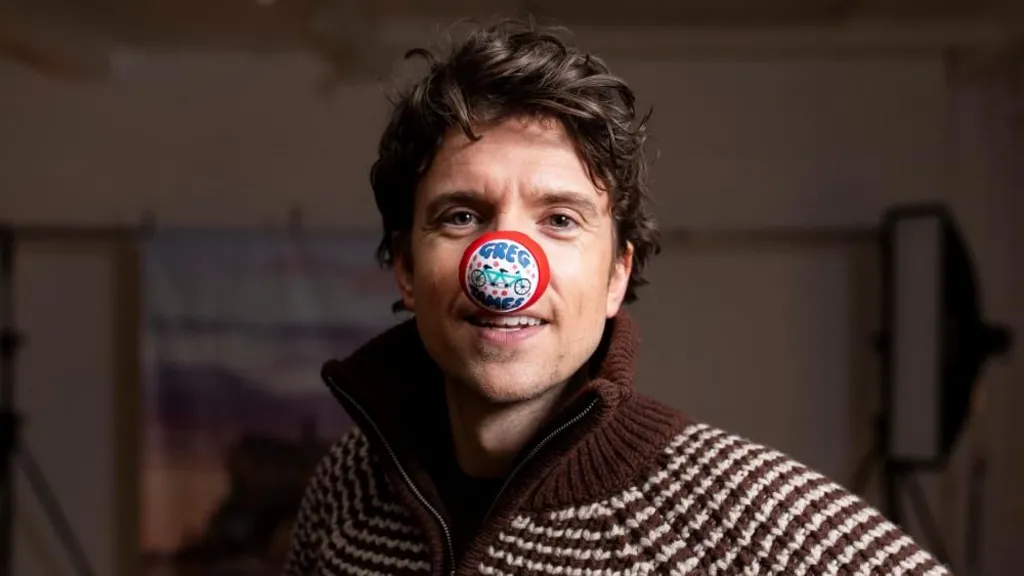 A close-up photo of Radio 1 DJ Greg James, he is wearing a brown and white stripped knitted jacket and has long curly brunette hair. He is staring directly at  the camera and has a red nose on his nose that has a picture of a tandem bike on it and says Greg James.