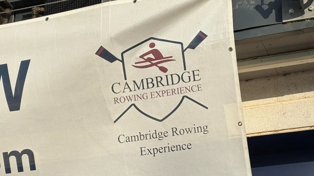 The corner of a white banner, which is hung on the outside of a rowing club's boathouse. This part of the banner shows a logo with the words "Cambridge Rowing Experience" written underneath a simple drawing of a single rower in a boat, pulling a stroke on their oars. The rower image is set inside a shield, behind which are two crossed oars. Below the logo is a repeat of the words "Cambridge Rowing Experience".  