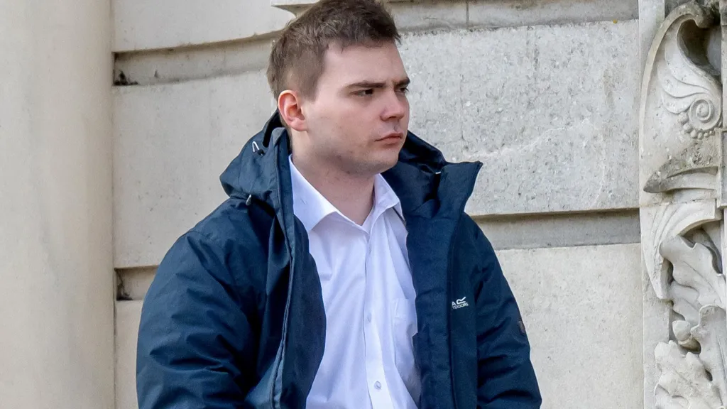 Oliver Beynon stands outside court in a white collared shirt and dark blue jacket, looking off to the side with a serious expression.