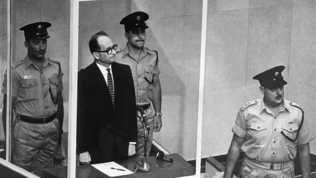 Adolf Eichmann at trial. He stands in a glass box surrounded by guards in an Israeli courtroom in the 1960s