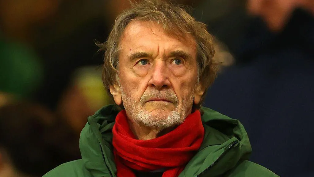 Jim Ratcliffe, with a grey stubbly beard, watches football in the stands wearing a red scarf and green jacket