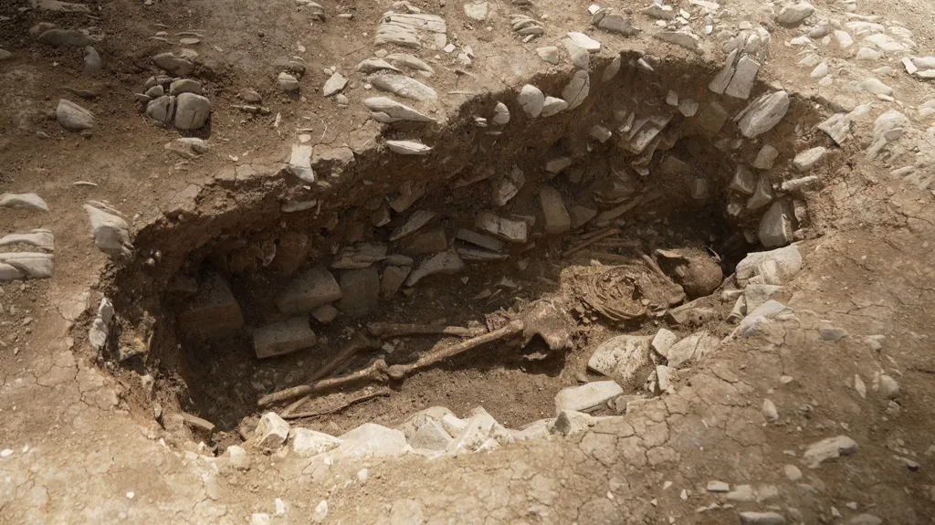 A human skeleton is lying in an oval-shaped grave cut into limestone bedrock. The bones are positioned naturally, with the skull at one end and legs extended. The ground surrounding the grave is light brown and cracked.