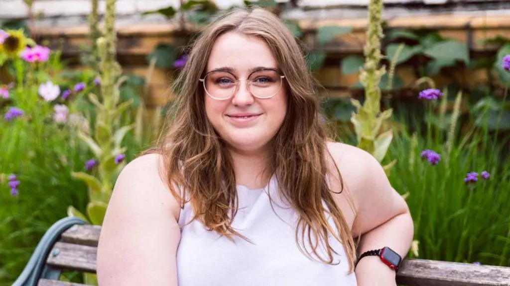 Cotton sitting on a bench with greenery in the background. She wears an apple watch and purple dress. She has long brown hair and wears thin, clear-framed glasses. It is a head and shoulders shot.
