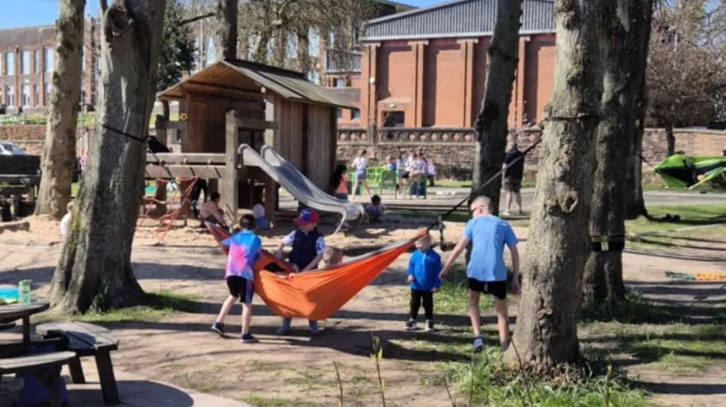 A sunlit playpark with parents and children playing among the trees