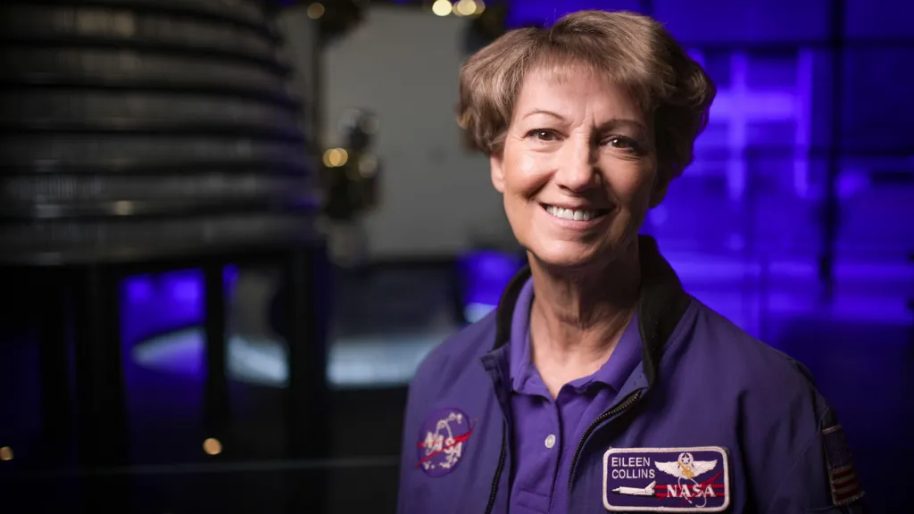 Eileen Collins wearing a blue jacket with a round Nasa patch on one side and a rectangular shuttle patch on the other with her name. She has short brown hair and brown eyes and is smiling as she looks directly to camera. She is in the space gallery at the Science Museum and an out of focus rocket engine can be seen behind her. 
