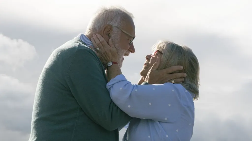 An elderly couple, both with grey hair. Holding each other's faces. The man is wearing a grey jumper and has a beard, the woman is wearing a blue polka dot top. They are gazing into each others eyes, with a sky pictured behind them.