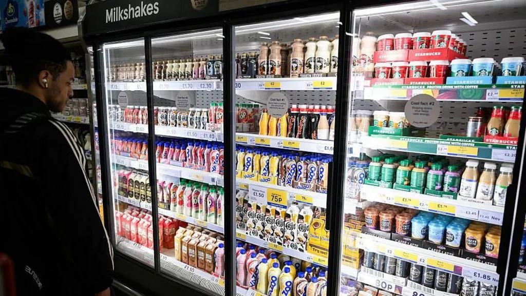A man with dark hair stands in front of a large supermarket fridge containing shelves of milk-based drinks, such as flavoured milkshakes and milky coffees like lattes, cappucinos and iced coffees.