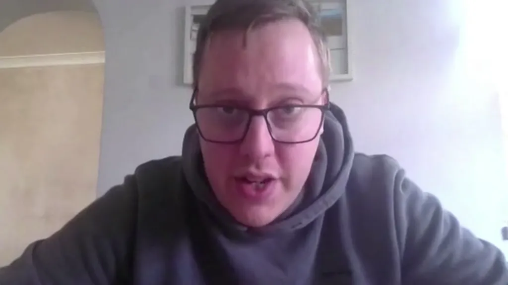A bespectacled man in a grey hoodie. He is being interviewed on Zoom and is responding to a question. He has his head slightly down and is looking at the camera.