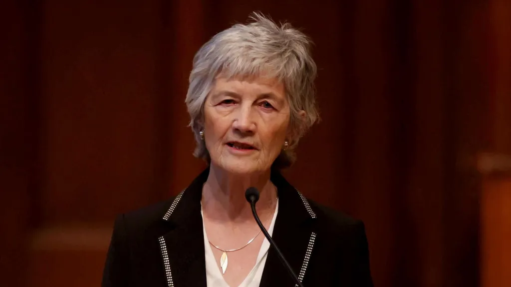 Shows a women with grey hair, a black jacket and white top, standing at a mic