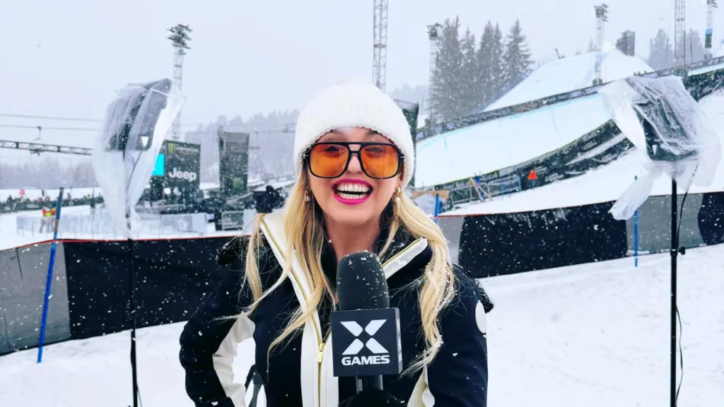 Aimee Fuller is wearing a white woollen hat, orange tinted glasses and a black coat in the snow holding a microphone. A slope set up can be seen behind her. 