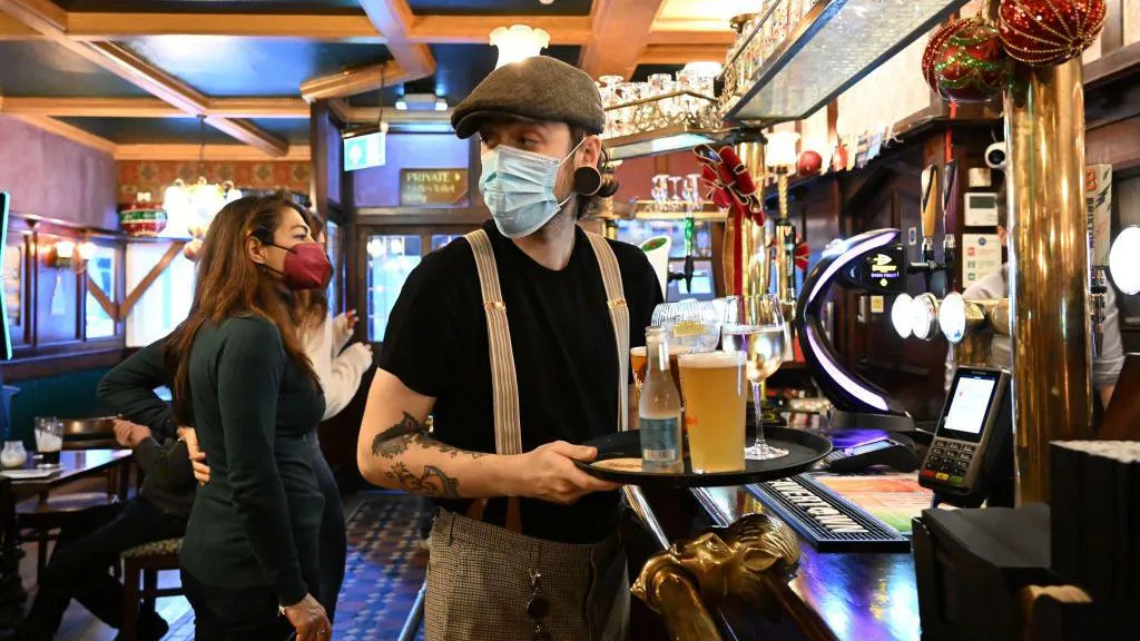 A bartender in vintage trousers, black T-shirt and a dark green flat cap wears a face mask as he carries a tray of drinks towards customers 