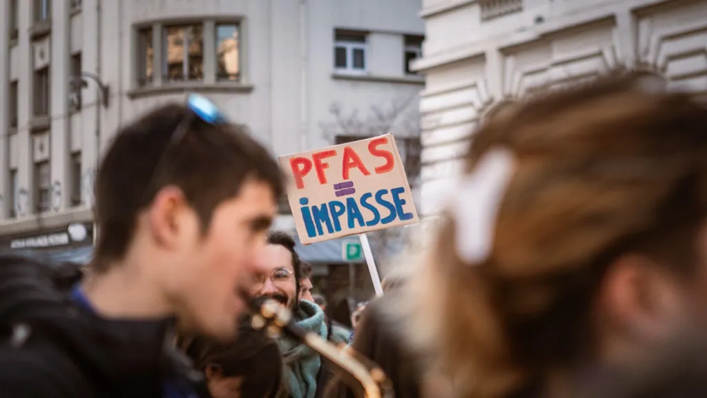 A protest image, with blurred faces in the foreground, and in the background a man smiling with a beard and glasses is holding a sign that reads "PFAS = IMPASSE"