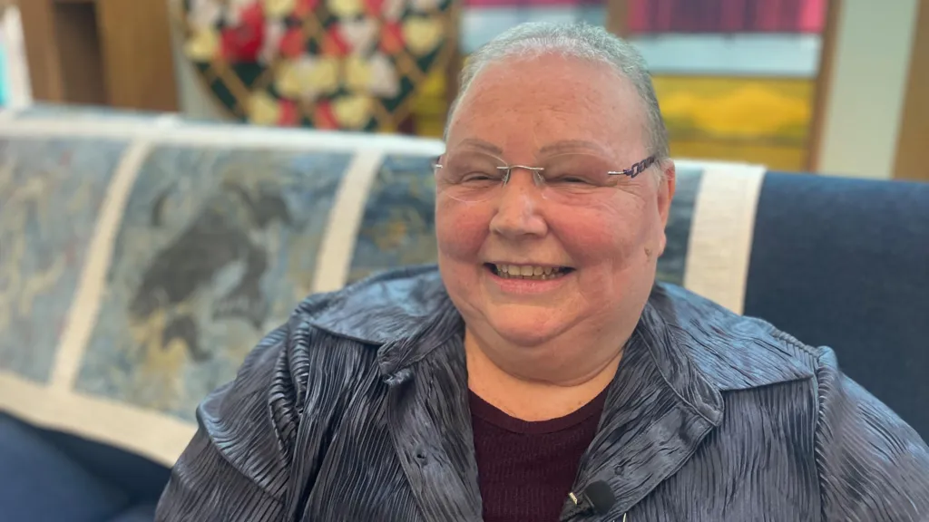 Karen sitting on a blue couch with a patterned throw over the back. She has short hair and glasses and is wearing a blue blouse and a burgundy top.