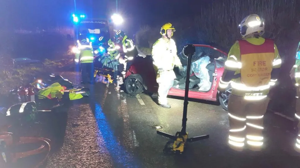 Image shows a scene of an accident. There is a red car, which belongs to Rebecca Rumens. The wheels are at an angle to the car and the doors are no long on the car. There are a number of emergency workers with yellow fluorescent clothing on. In the background, there is an ambulance with it's blue lights on. Emergency workers are tending to someone who is led on their backs on a stretcher. To the left of the picture, there is lots of kit piled along the road. In the middle of the picture, there is a standing light used to help the workers see what they are doing as it is night time. 
