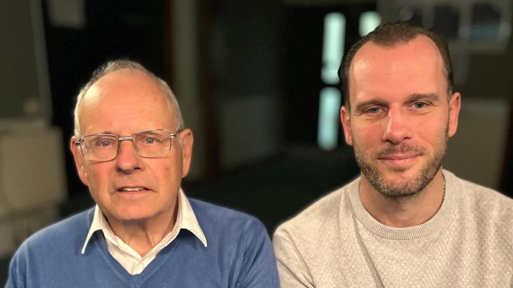 Graham Hall and Steven Hall sit next to each other and smile at the camera. Graham has thin grey hair and wears a blue jumper with a white shirt underneath. Steven has short dark hair and is wearing a cream coloured jumper.