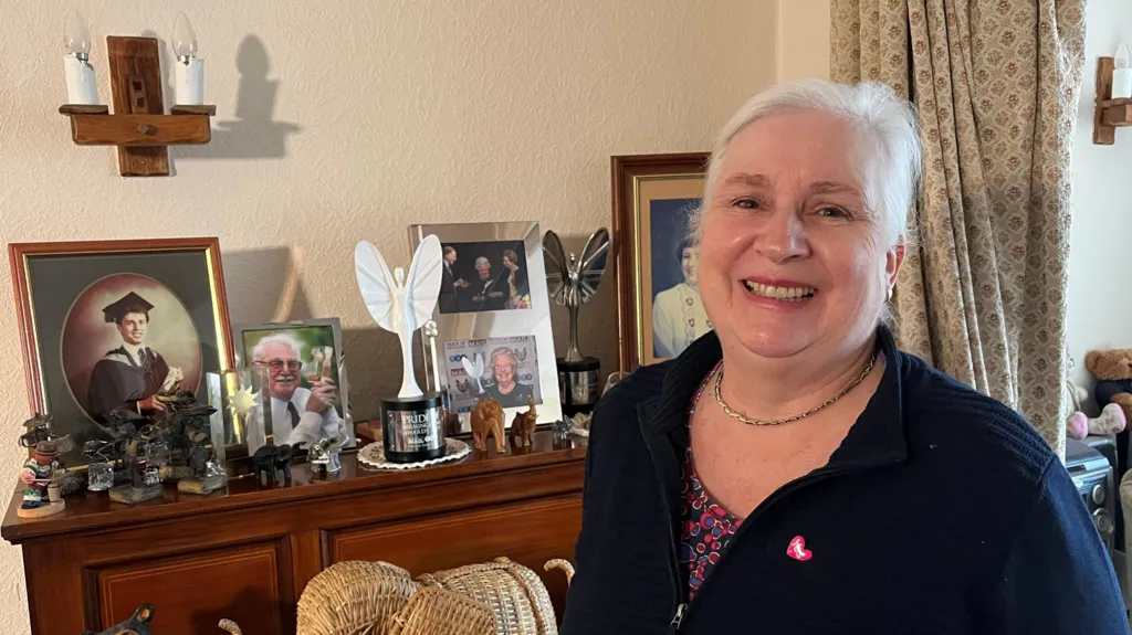 A woman with silver hair smiles at the camera. She stands in a living room in front of a wooden cabinet with lots of family photos and an award on it.