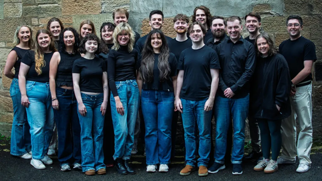 A group of 19 young people in various black tops and blue jeans, stand in front of a stone wall. They are all smiling. 