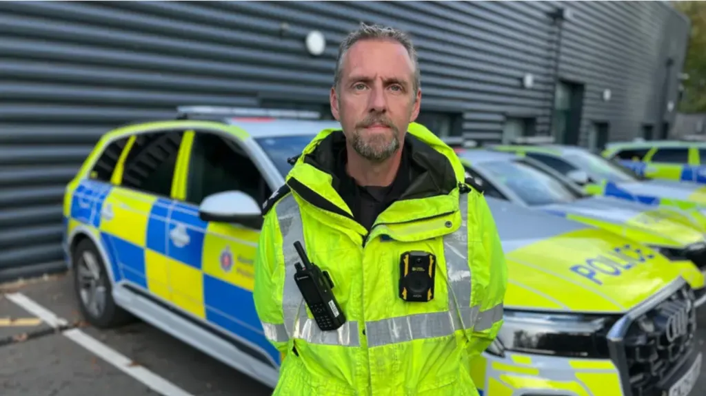 Det Sgt Chris Wade is wearing a high-vis policing jacket, with his radio and body cam. He is standing in front of a row of four police cars. 