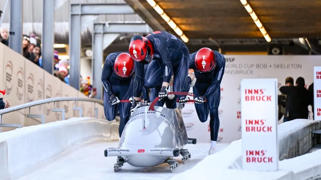 A four-man Trinidad and Tobago bobsleigh team in dark blue suits and red “T&T” helmets pushes a silver sled marked “LotteryWorld” at the start line of an IBSF World Cup race in Innsbruck. The athletes lean forward in unison on the icy track, with spectators watching from the side and event signage visible in the background.