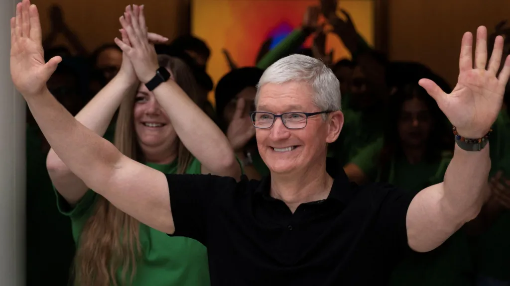 Apple CEO Tim Cook, in a black polo and smiling wide as he waves with both hands in the air at a crowd outside of a new Apple retail store. 