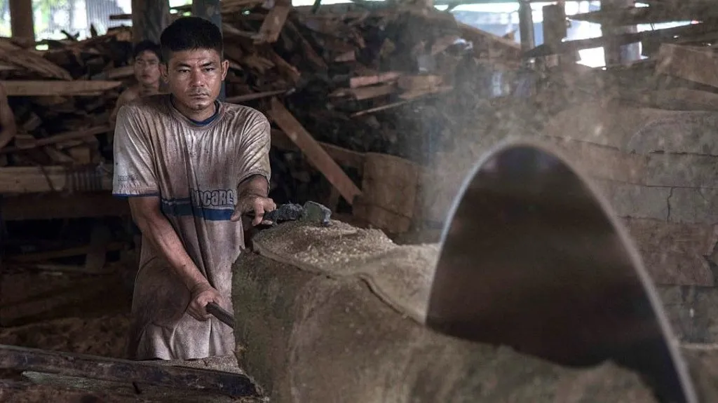 A large spinning blade cuts through a log of wood with a worker, covered in sawdust, controlling the process at the back of the shot. 
