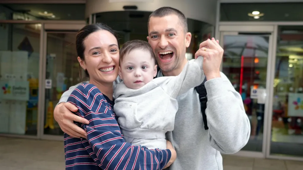 A slim woman with dark hair tied back, wearing a navy blue and red stripe top and a dark haired man in a grey hoodie. They stand together, holding a dark haired male toddler, both smiling. Callum holds up the boy's arm as though in celebration.