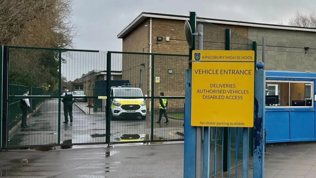 A police van is parked inside the gates of Kingsbury High School in Brent, while officers stand nearby on a wet road with school buildings and signage visible.