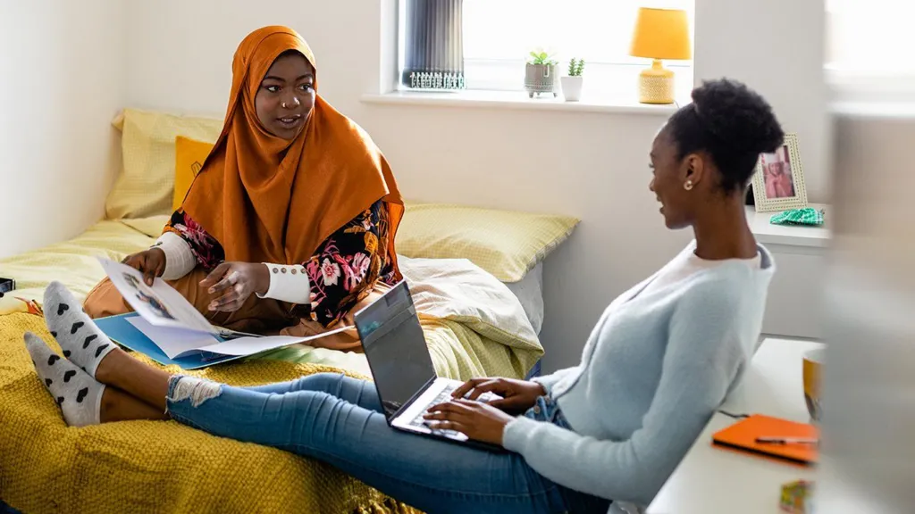 Two female university students chat in a student dorm room. One sits on the bed flicking through notes inside a blue ring binder, while the other is typing on a laptop resting on her lap.
