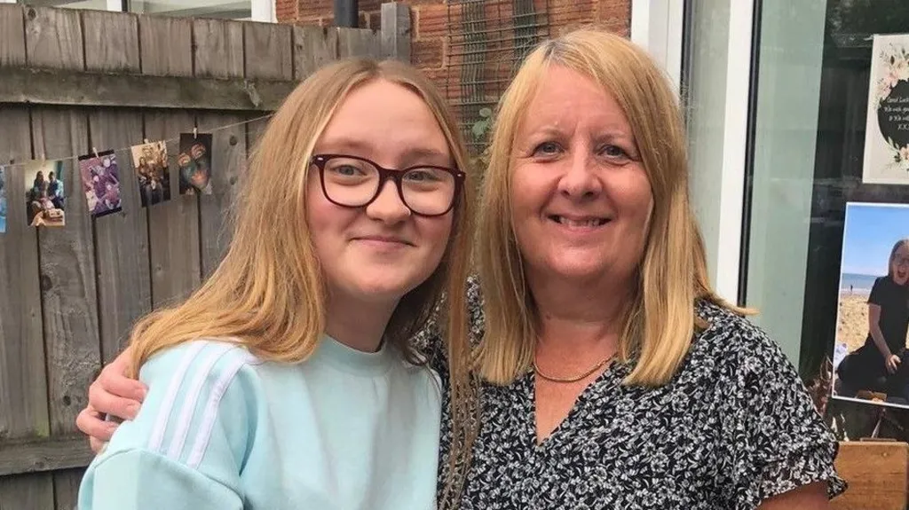 A daughter and mother standing arm in arm in a back garden, with a wooden fence, patio door and various colourful photos in the window and hanging from the fence. The daughter, a young woman, has long strawberry blond hair and wears purple-framed glasses and a light blue jumper with white stripes on the shoulder. The mother has similar hair and wears a black floral blouse, with a thin gold necklace around her neck.