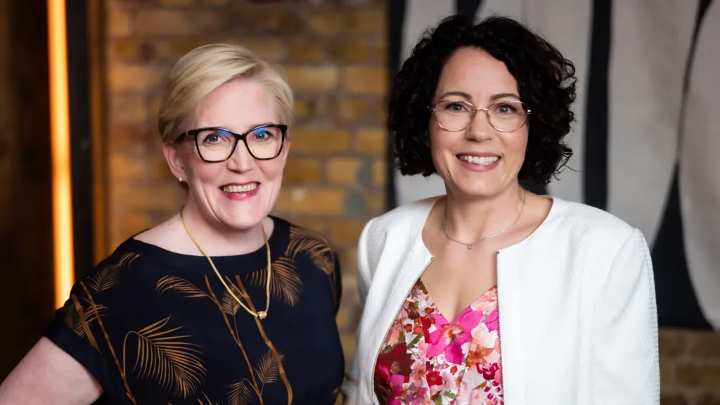 With blonde hair and wearing a black blouse Monika Liikamaa  stands next to Denise Johansson who wears a white jacket and flowery pink blouse. 