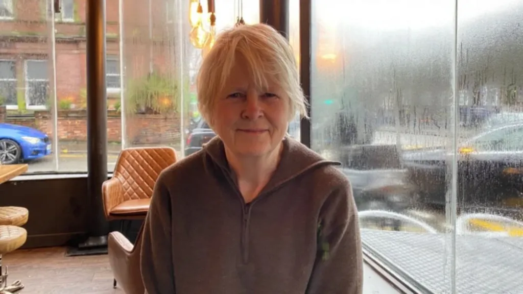 Williams is sat on a chair inside a cafe next to a large glass window which has condensation on it. She is wearing a brown top and has short blonde hair. There are empty, orange chairs behind her and the street can be seen outside