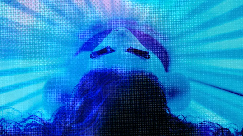 A woman lies on her back in a sunbed, wearing goggles - tubes surround her emitting blue light.