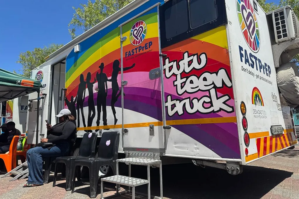 A rainbow striped mobile clinic is parked up. On the side it says 'tut teen truck'. There are some black chairs in front - a woman checking her phone is sitting on one of them.