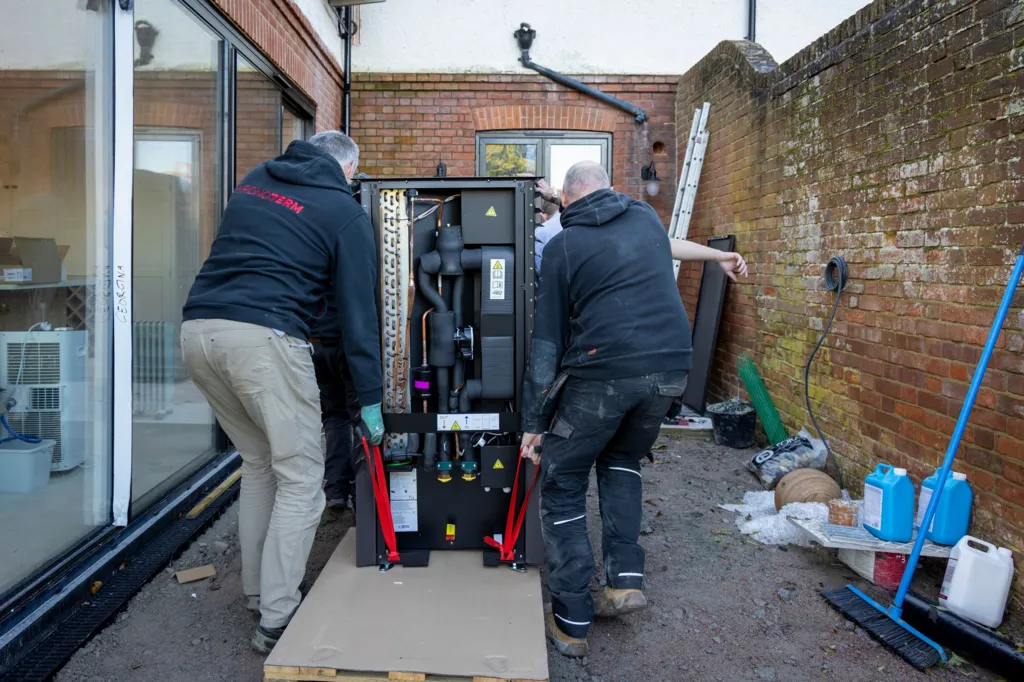 Three men with their backs to the camera are shown lifting a black heat pump on a trolley towards the back of the house. The men wear dark work clothes and boots. The house is red brick with a long glass door to the left and the property boundary wall to the right. Against the wall sits work tools and a blue broom. 