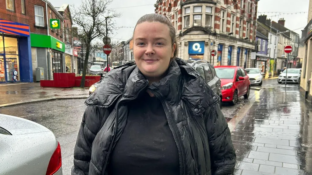 A woman stands close to the camera on a very rainy day. She has brown hair tied back in a ponytail. She is wearing a black puffer coat. The pavement is slick and reflective in the rain behind the woman, backed also by a line of traffic. Behind her is a busy street in strabane, with buildings like a subway and o2 shop in vision. 