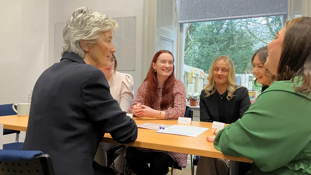 Shows an older woman with grey hair and a dark jacket talking to five younger women
