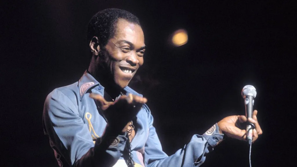 Nigerian singer Fela Kuti, in a pale blue long-sleeved shirt with bits of yellow and pink embroidery, smiles and holds up his hands towards the audience at Vredenburg in Utrecht, Netherlands on 3 November 1988