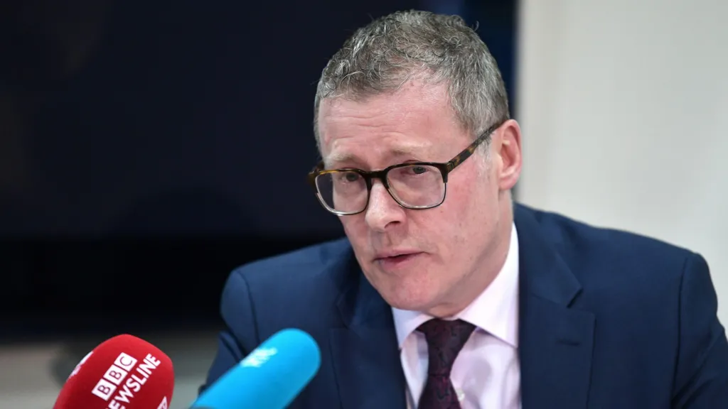 Kevin Winters, a man with short, greying hair and glasses, leans towards microphones during a press conference in 2024.  He is wearing a navy suit, a pink shirt and a dark red tie. 