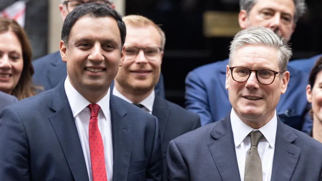 Two smiling men stand side-by-side posing for a photograph. They are both wearing dark suits, white shirts with ties. 