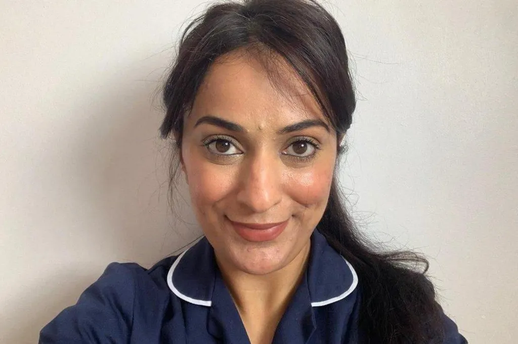 A selfie taken by a nurse wearing blue scrubs. She looks at the camera and smiles, while standing in front of a white background.