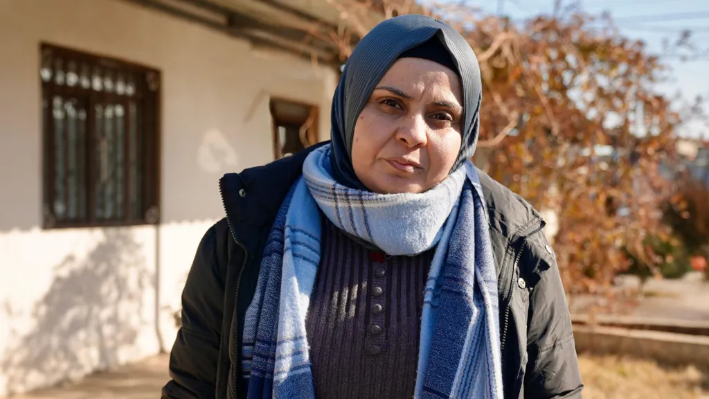 Hekmiya Ibrahim stares into the camera. She is in a grey headscarf and a black puffer jacket, wearing also a light blue striped scarf for warmth
